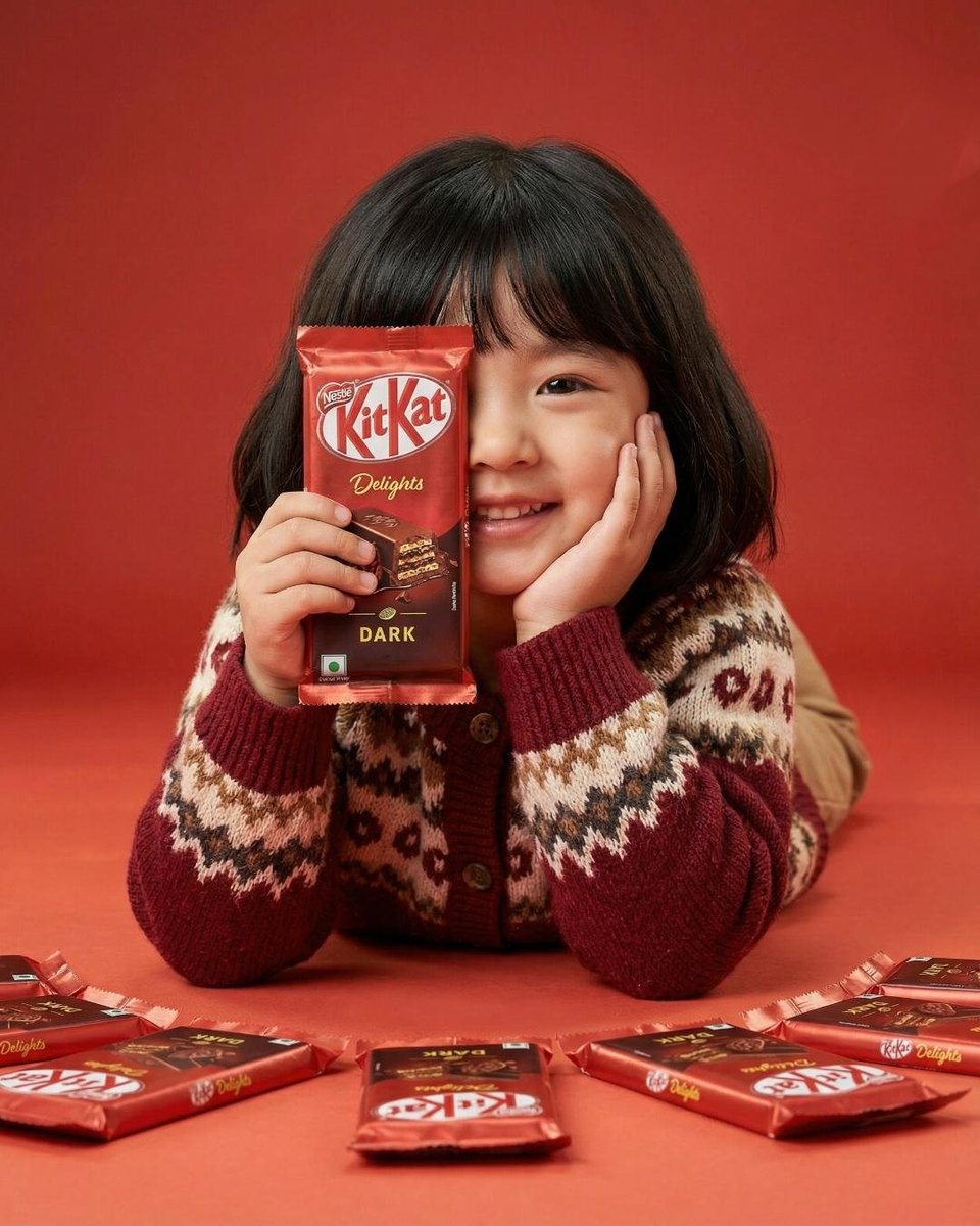 A cute young girl with short black hair and bangs smiling while holding a red KitKat Dark chocolate…